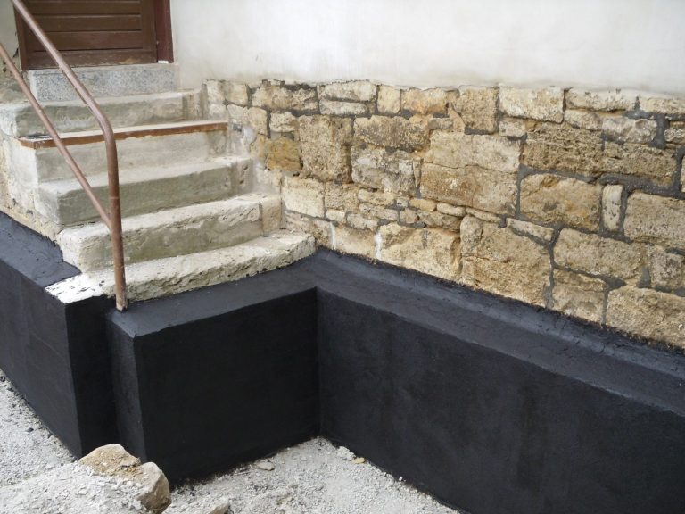 Der Sockel eines Hauses wurde mit Beton und Bitumen gegen Feuchtigkeit abgedichtet. Eine Sandsteintreppe führt von hinten ins Haus.